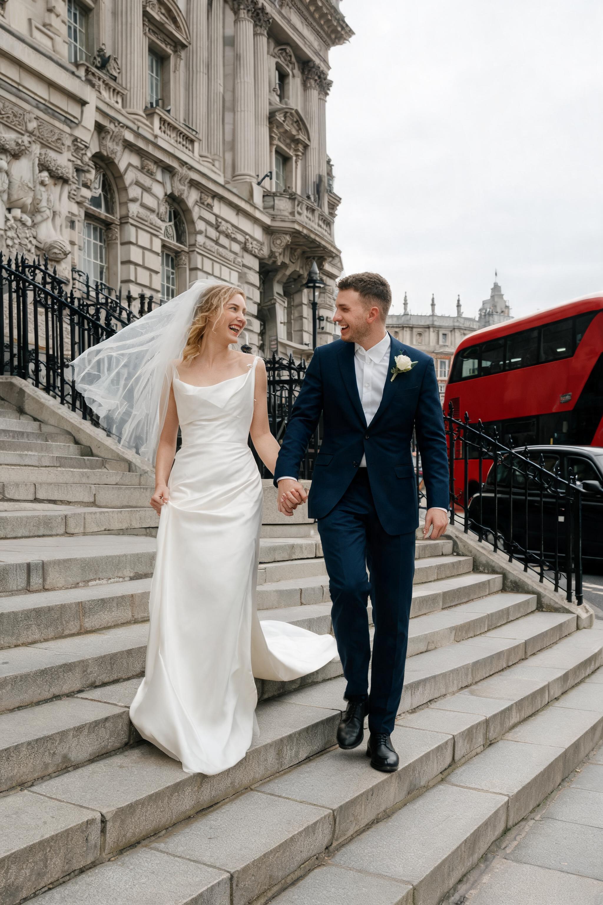 Bride and groom descending historic London city hall steps, she in ivory silk gown, he in navy suit, with red bus and black c