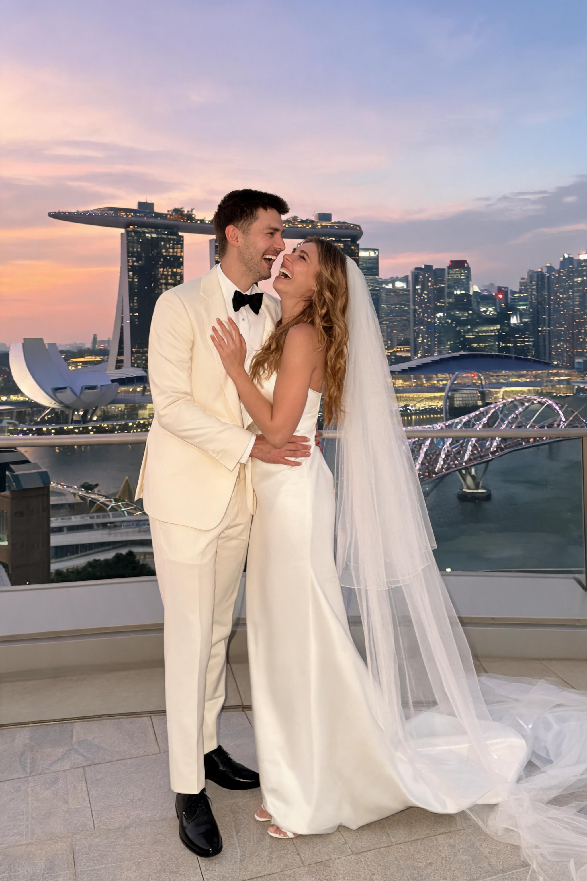 Couple in wedding attire on Marina Bay Sands rooftop at dusk, laughing, with Singapore skyline and city lights behind them.